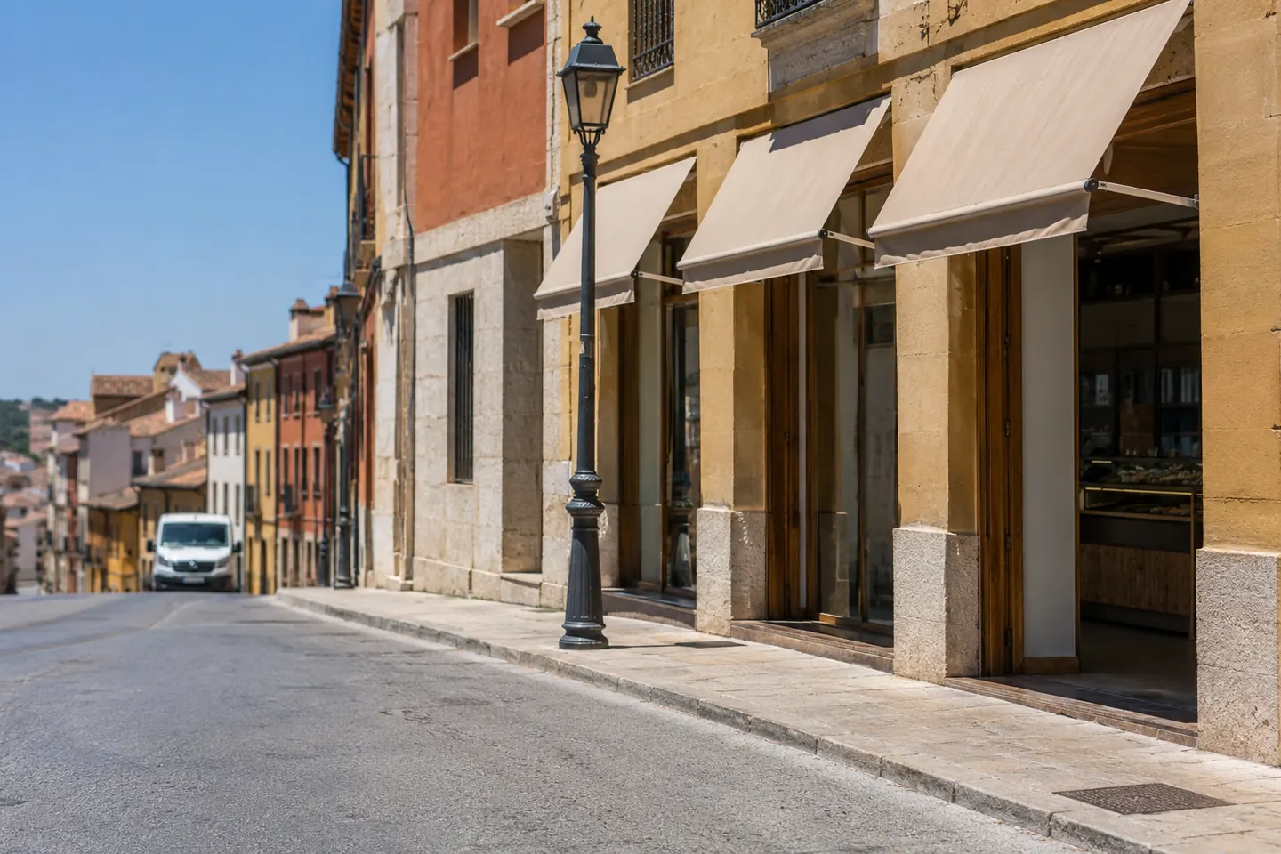 Calle comercial en cuesta con varios locales de persiana subida y toldos, a media mañana